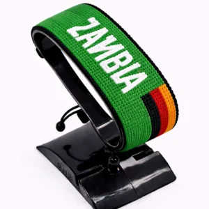 Handcrafted Zambia name flag bracelet made with green, red, black, and orange beads, featuring the name “Zambia,” adjustable beaded wrist accessory, displayed on a black stand.